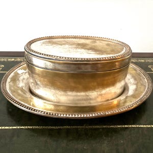 May include: A silver-toned, oval box with a matching saucer. The box has a lid and sits on the saucer. Both pieces have a decorative beaded edge. The set is displayed on a dark green surface.