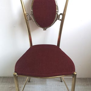 May include: A gold metal chair with a burgundy velvet seat and a decorative back. The chair has a tall, ornate back with a scroll design and a central oval.