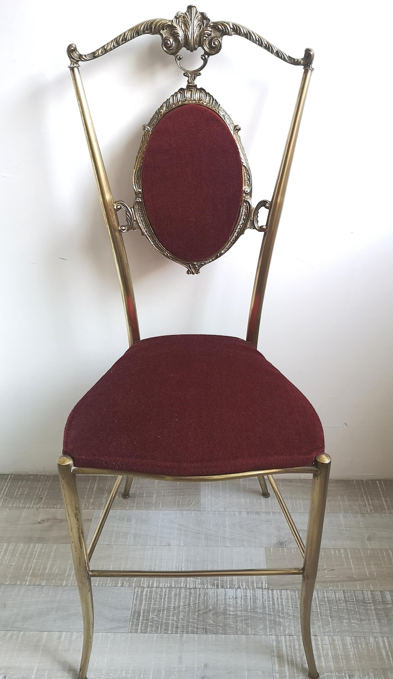 May include: A gold metal chair with a burgundy velvet seat and a decorative back. The chair has a tall, ornate back with a scroll design and a central oval.