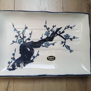 May include: White rectangular serving dish with a blue and black floral design. The dish has a raised rim and a blue border. The floral design features a stylized tree with blue flowers and black branches. The dish has a gold label with Japanese text.