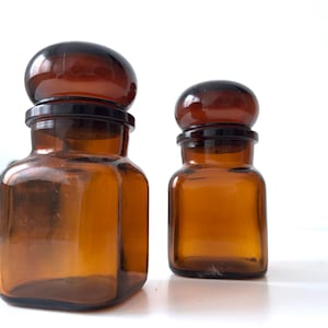 Puede incluir: Dos frascos de boticario vintage de vidrio ámbar con tapones marrón oscuro. Los frascos tienen una base cuadrada y hombros redondeados. Los tapones son redondos y se asientan sobre un borde negro. Los frascos son de diferentes tamaños.