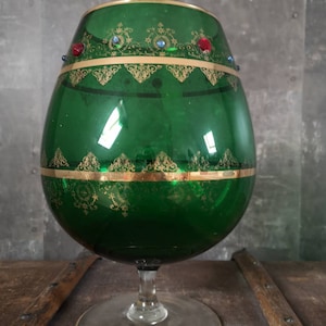 May include: A green glass vase with gold trim and colorful jewels around the rim. The vase has a round body and a clear glass stem.