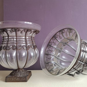 May include: Two clear glass vases with a decorative wire design. The vases have a round base and a wide opening. The wire design is intricate and adds a touch of elegance to the vases.