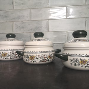 Op de afbeelding: Drie witte keramische potten met groene deksels en bloemmotieven. De potten hebben zwarte handvatten en zijn in een rij op een grijs oppervlak gerangschikt.