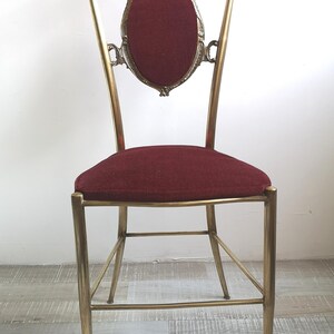 May include: A gold metal chair with a burgundy velvet seat and back. The chair has a decorative, ornate design with a curved back and a round, framed back panel.
