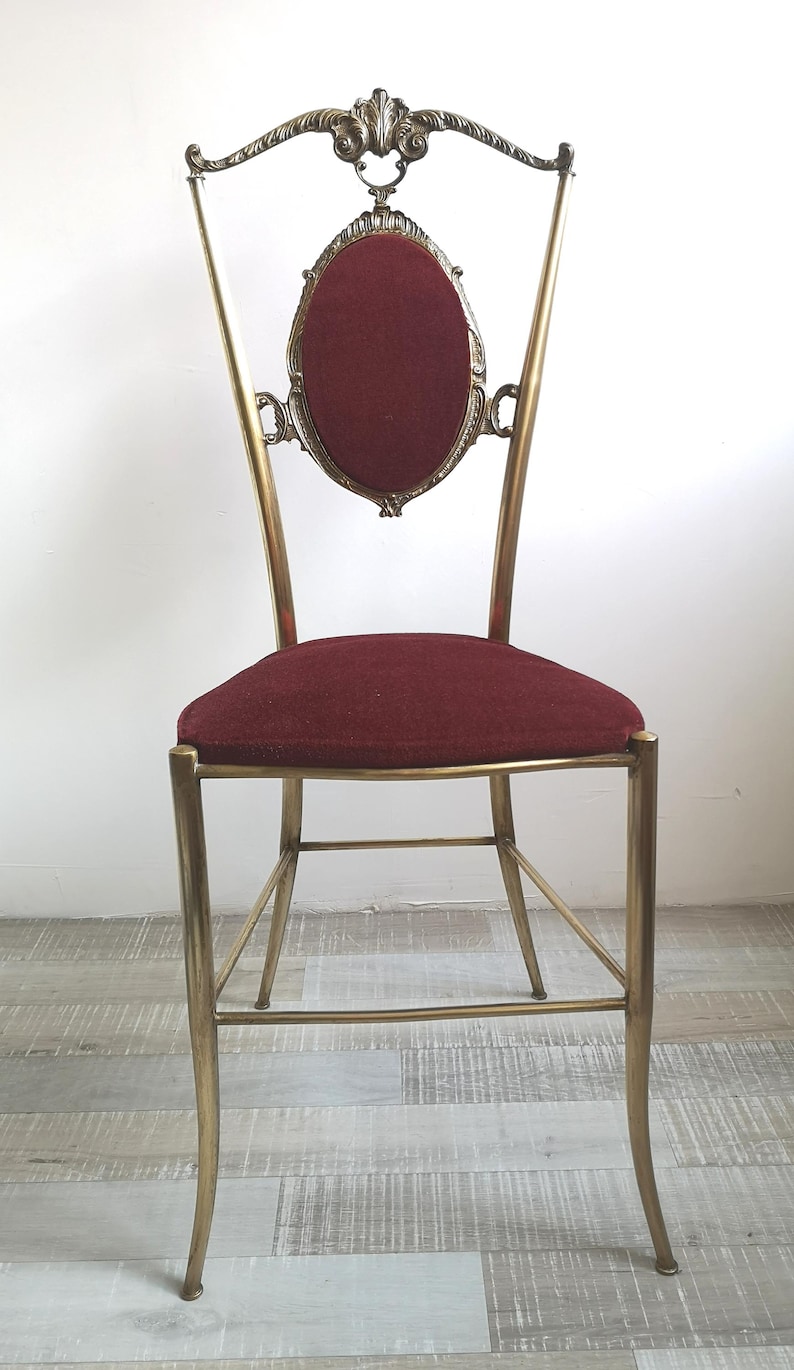 May include: A gold metal chair with a burgundy velvet seat and back. The chair has a decorative, ornate design with a curved back and a round, framed back panel.