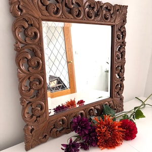 May include: Square wooden mirror with an ornate, carved frame. The frame is a warm brown color and features intricate floral and scrollwork designs. A bouquet of red, orange, and purple flowers rests in front of the mirror.