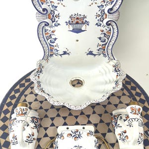 Op de afbeelding: Een witte keramische wandwastafel met een geschulpte rand, versierd met blauwe en oranje bloemmotieven. De set bevat bijpassende zeepdispensers, een handdoekring en een decoratieve backsplash, alles op een mozaïek tegel tafel.