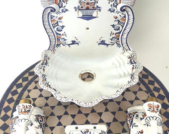 Op de afbeelding: Een witte keramische wandwastafel met een geschulpte rand, versierd met blauwe en oranje bloemmotieven. De set bevat bijpassende zeepdispensers, een handdoekring en een decoratieve backsplash, alles op een mozaïek tegel tafel.