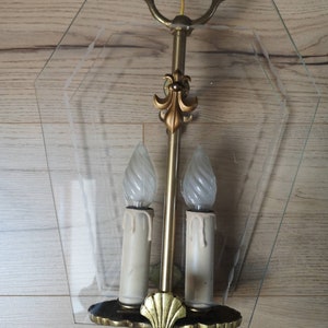 May include: A gold-toned, two-light wall sconce with a clear glass shade. The sconce has a decorative fleur-de-lis design and a scalloped bottom.