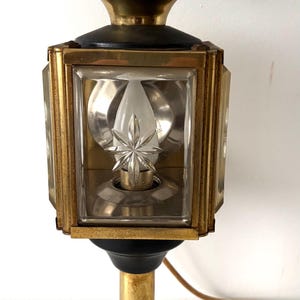 Puede incluir: Una lámpara tipo linterna de latón con una carcasa de vidrio cuadrada y un detalle de cristal en forma de estrella en su interior. La lámpara tiene una parte superior redondeada de latón, una base negra y un mango cilíndrico de latón.