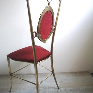 May include: A tall, ornate brass chair with a red velvet seat and back. The back features a large oval frame with a red velvet insert.