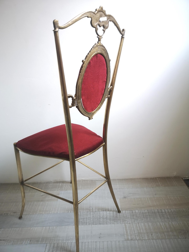 May include: A tall, ornate brass chair with a red velvet seat and back. The back features a large oval frame with a red velvet insert.