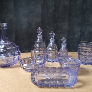 Op de afbeelding: Een verzameling vintage toiletartikelen van lavendelkleurig glas. De set bevat een grote fles, verschillende kleinere flessen met stoppen, een ringschaal, een afgedekte schaal, een rechthoekige lade en een beker. Het glas heeft een gestructureerd, geometrisch ontwerp.