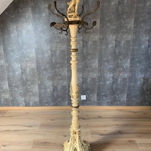 Op de afbeelding: Een witte houten kapstok met een gesneden cherubfiguur bovenop. De kapstok heeft meerdere haken en een decoratieve basis.