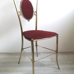 May include: A tall, ornate chair with a gold metal frame and a red velvet seat. The chair has a decorative back with a scroll design and a small oval-shaped cushion.