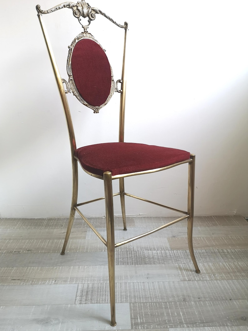 May include: A tall, ornate chair with a gold metal frame and a red velvet seat. The chair has a decorative back with a scroll design and a small oval-shaped cushion.