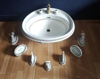 Op de afbeelding: Witte porseleinen badkamer wastafel met gouden rand en decoratieve accenten. De wastafel is omgeven door bijpassende wandgemonteerde zeepdispensers en handdoekhaken.