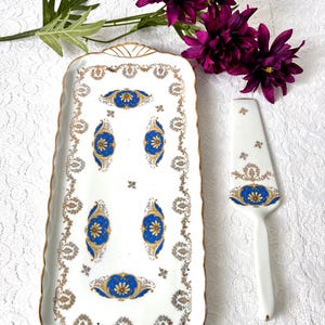 Könnte beinhalten: Ein rechteckiges Serviertablett aus weißem Porzellan und ein passender Tortenheber mit gewelltem Rand und Goldverzierung. Das Set ist mit kunstvollen Goldfiligranmustern und leuchtend blauen Blumenmedaillons auf der weißen Oberfläche verziert.