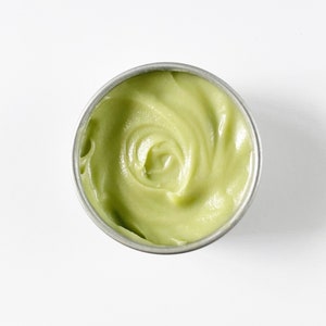 May include: A small, round, silver container filled with a light green, smooth, creamy substance.