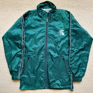 Puede incluir: Una chaqueta cortavientos verde con ribetes blancos y una cremallera negra. La chaqueta tiene un logotipo de Spartan en el pecho izquierdo.