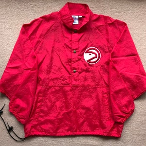 May include: Red windbreaker jacket with a white Atlanta Hawks logo on the front. The jacket has a snap button closure and a kangaroo pocket.
