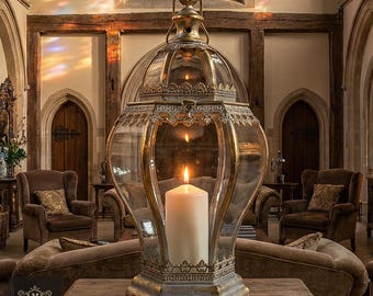 May include: A large, ornate glass and metal lantern with a lit candle inside. The lantern has a gold-colored frame and a clear glass enclosure. Dimensions: 17.7 inches high, 9.8 inches wide, and 9.8 inches deep. Weight: 4.6 pounds. The lantern sits on a wooden table.