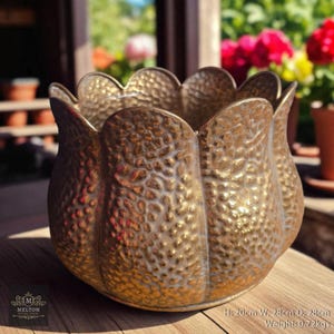 May include: A gold-coloured, textured metal planter with a scalloped rim. The planter is 20cm high, 28cm wide, and 28cm deep, and weighs 0.72kg. The planter has a hammered texture and is sitting on a wooden surface. The image includes the text "MELTON".
