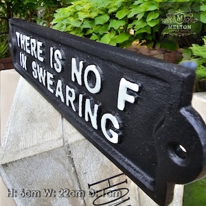 May include: A black cast iron sign with the text "THERE IS NO F IN SWEARING" in white lettering. The sign is 22cm wide, 6cm high, and 1cm deep. The sign has a decorative end with a hole for hanging.
