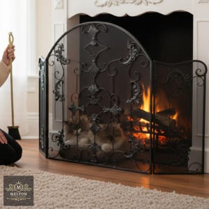 May include: An elaborate black metal fireplace screen with a decorative scroll pattern. The screen is positioned in front of a fireplace with a burning fire. A person is holding a brass fireplace tool. The Melton Cottage Online logo is visible.