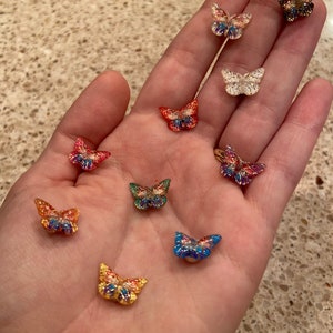 May include: A collection of small, colorful butterfly-shaped embellishments. The butterflies feature a variety of colors, including red, blue, gold, and green, with glitter accents. These decorative elements are ideal for crafts and DIY projects.
