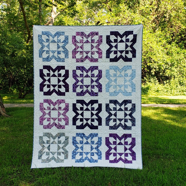 Quilt with blue and purple starburst bloom blocks on a light background.  Quilt is photographed outside on grass with trees in the background.