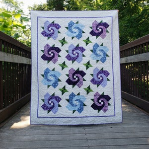 Quilt with 12 swirling bloom blocks in purple and blue and 6 green 4-ppointed star cornerstones, all on a cream background.  Quilt is on a bridge with a weathered wood deck and rusty metal railings.