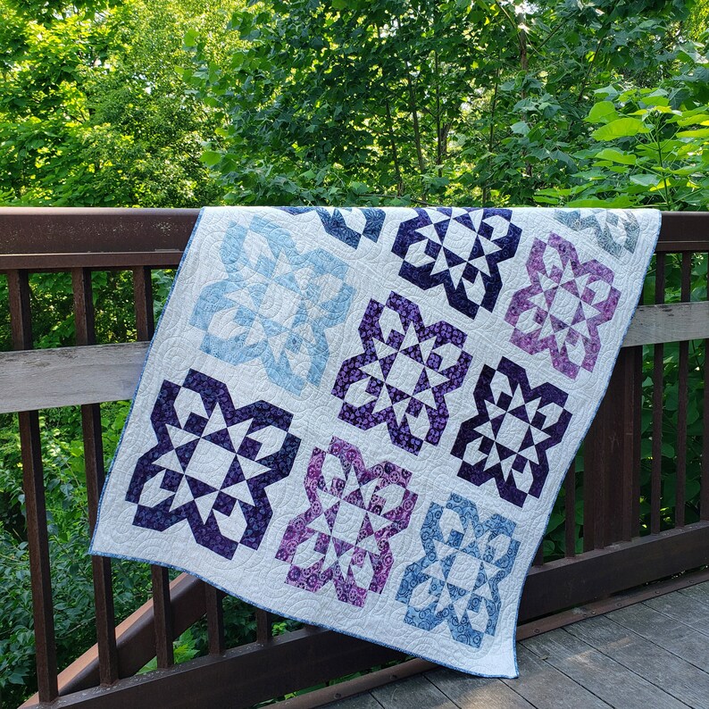 Quilt with blue and purple starburst bloom blocks on a light background.  Quilt is draped at an angle over a rusty railing against leafy greenery.