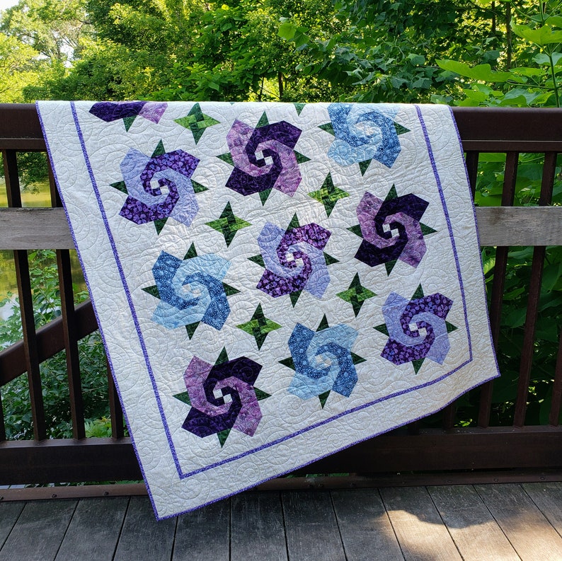 Quilt with 12 swirling bloom blocks in purple and blue and 6 green 4-pointed star cornerstones, all on a cream background.  Quilt is draped on a rusty metal railing with greenery in the background.
