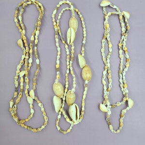 May include: Three seashell necklaces with varying designs. The necklaces feature a mix of small, light-colored shells and larger, cream-colored cowrie shells. The shells are strung together, creating a natural, beachy aesthetic. The background is a solid purple.