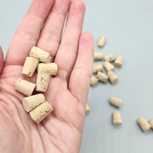 May include: A collection of small, natural cork stoppers. The corks are cylindrical with a tapered shape, showing a light brown color and visible grain. Some stoppers are held in a hand, while others are scattered on a light gray surface.