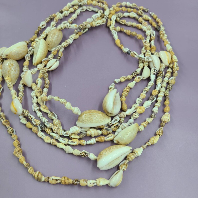 May include: A multi-strand necklace composed of various seashells in shades of cream, tan, and brown. The necklace features a mix of small, round shells and larger, oval-shaped shells, creating a layered effect. The background is a solid purple.