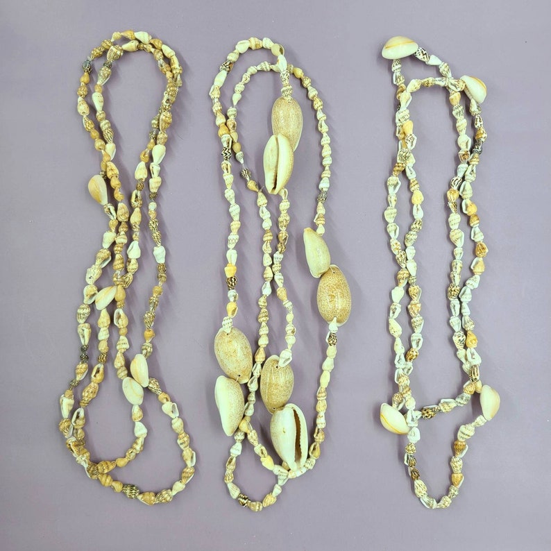 May include: Three seashell necklaces with varying designs. The necklaces feature a mix of small, light-colored shells and larger, cream-colored cowrie shells. The shells are strung together, creating a natural, beachy aesthetic. The background is a solid purple.