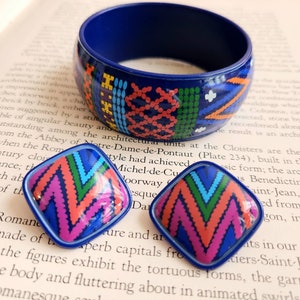 Könnte beinhalten: Ein blaues Armreif-Armband und passende quadratische Ohrringe. Der Schmuck weist ein farbenfrohes geometrisches Muster in Rot, Orange, Grün und Lila auf. Die Ohrringe und das Armband werden auf einer Buchseite präsentiert.