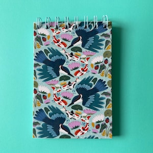 May include: A spiral-bound notepad featuring a repeating pattern of blue birds, koi fish, and lotus flowers on a light blue background. The design includes red, white, and pink accents, creating a vibrant and nature-inspired aesthetic.