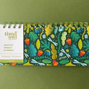 May include: A desktop weekly planner with a spiral binding. The cover features a dark green background with a colorful pattern of leaves, acorns, dragonflies, and flowers. The planner includes the text "Tiny Owl Stationery".