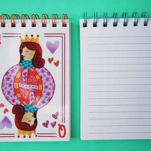 May include: Two spiral-bound notebooks. One features a playing card design with a queen illustration, hearts, and the letter "Q". The other notebook has lined pages. The notebooks have copper and blue colored spiral bindings.
