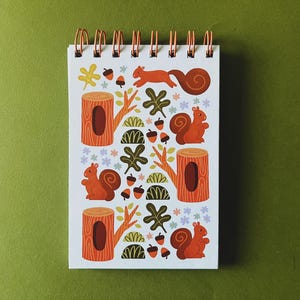 May include: A spiral-bound notepad with a whimsical forest-themed design. The illustration features orange squirrels, tree stumps, acorns, and foliage in shades of brown, green, and yellow. The notepad has a white background and copper-coloured spiral binding.