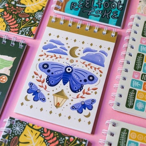 May include: A set of six colourful spiral bound notebooks with various designs. One notebook features a blue butterfly with an eye design, another has a repeating pattern of squares with different colours and shapes, and another has a floral pattern with green leaves and pink flowers.