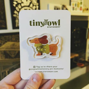 May include: A clear acrylic pin featuring a dachshund dog with brown fur, a brown ear, and a green sweater with yellow spots. The pin is on a white card with the text "tiny owl stationery". The card also includes the text "Tag us to share your @tinyowlstationery pin moments! tinyowlstationery.com".