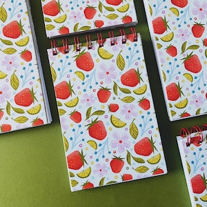 May include: A collection of spiral-bound notebooks with a repeating pattern of red strawberries, yellow lemon slices, pink flowers, and green leaves on a white background. The notebooks have red wire binding.