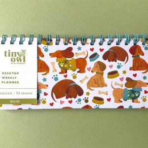 May include: A desktop weekly planner with a dachshund dog pattern. The planner features a white cover with the text "tiny owl stationery" and "desktop weekly planner". The spiral binding is teal. The background is a light green.