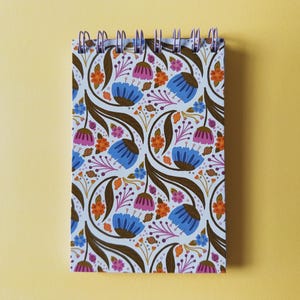 May include: A spiral-bound notebook with a floral pattern. The design features blue, pink, and orange flowers with brown stems and leaves on a white background. The notebook is on a yellow surface.