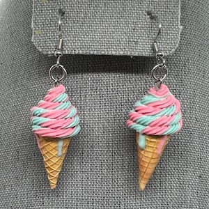 Op de afbeelding: Een paar bungelende oorbellen met miniatuur ijsjes. Elke kegel heeft een wafeltextuur, bekroond met roze en turquoise gedraaid "ijs" en een lichtblauwe druppel. De oorbellen hangen aan zilverkleurige haken.
